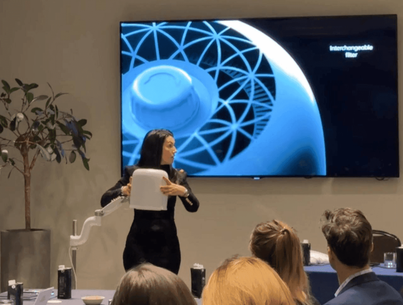 A woman presents a white square object in front of a large screen showing a blue geometric design.