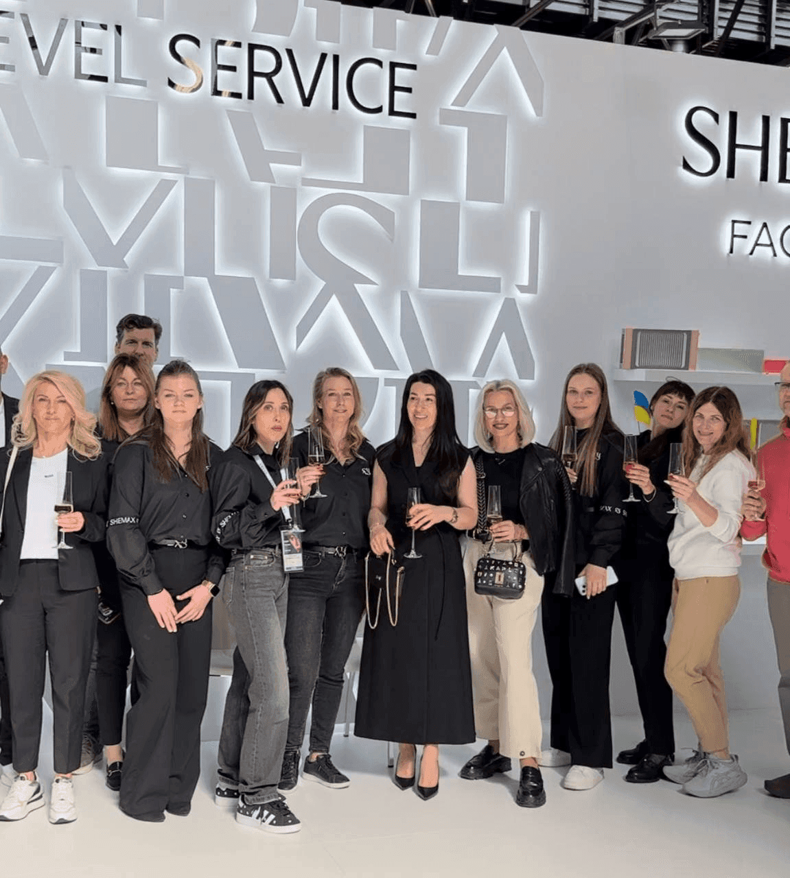 A group of people stand together in front of a SHEWAX branded wall at an event.