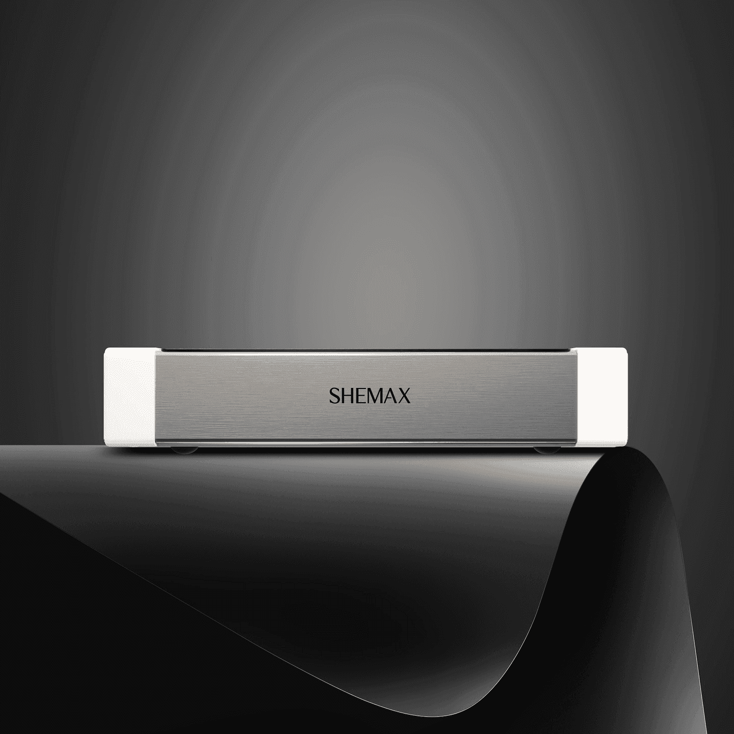 A gray SHEMAX device with white ends sits on a reflective surface against a dark background.