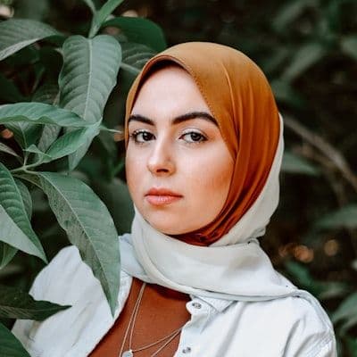 A woman wearing a hijab and white shirt stands near green foliage.