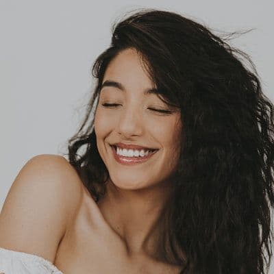 A woman with long, dark curly hair closes her eyes and smiles.