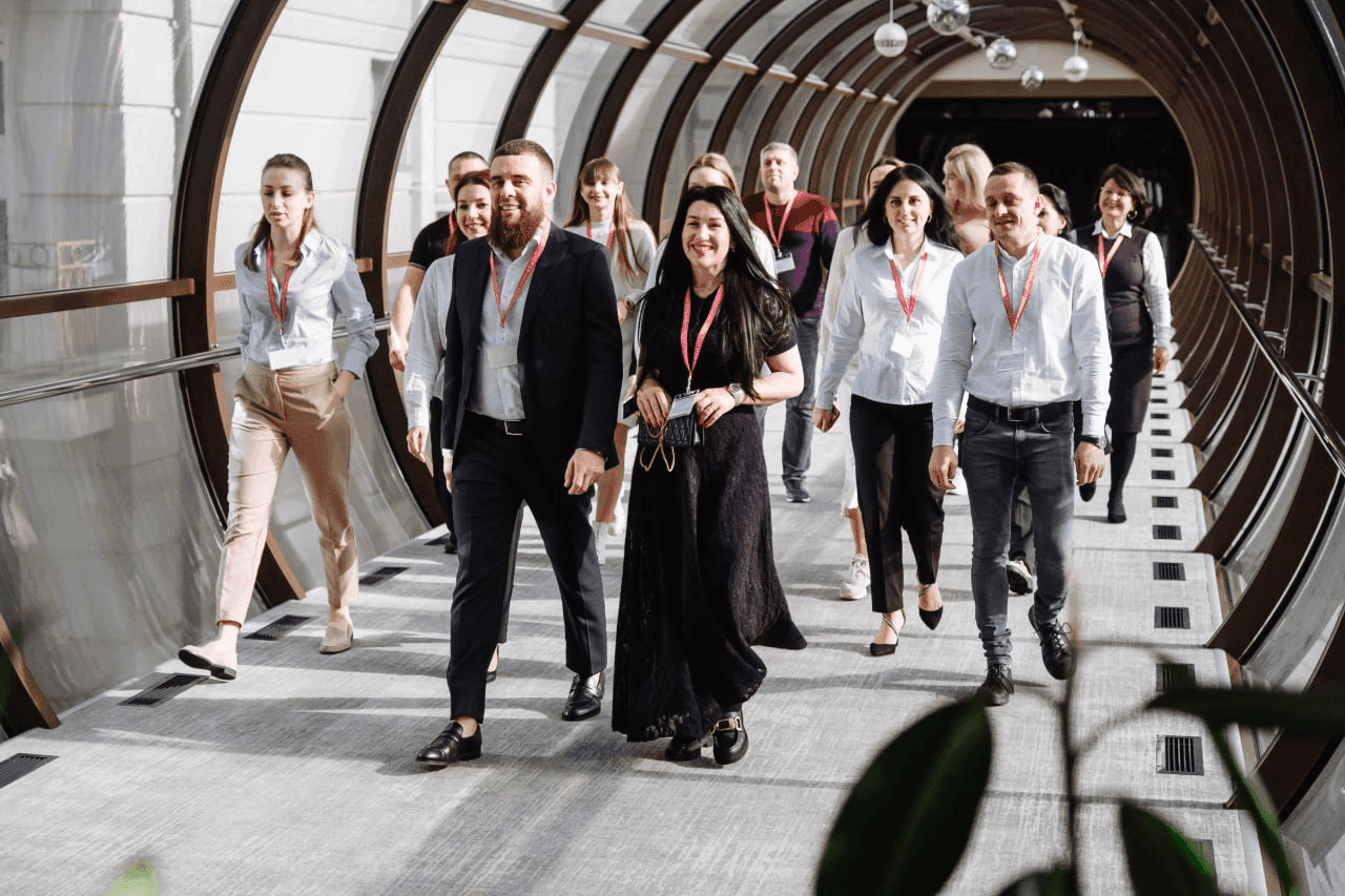 A diverse group of people walks down a modern, sunlit corridor, all wearing lanyards.
